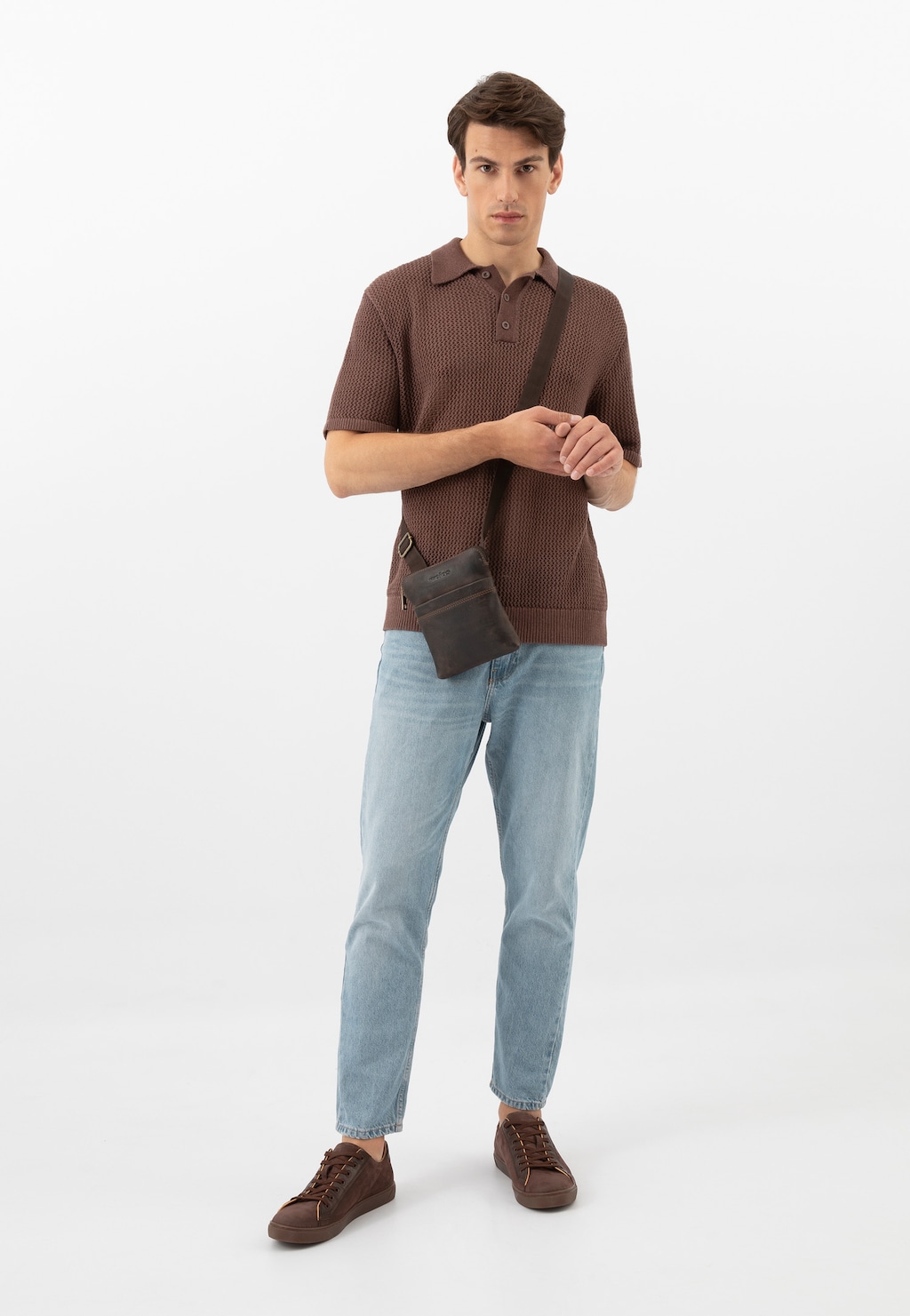 Model w brązowej koszulce polo i jasnych jeansach, prezentujący ciemnobrązowe trampki męskie Wojas model 10227-72 ze skórzanymi wstawkami, z małą torbą na ramieniu.