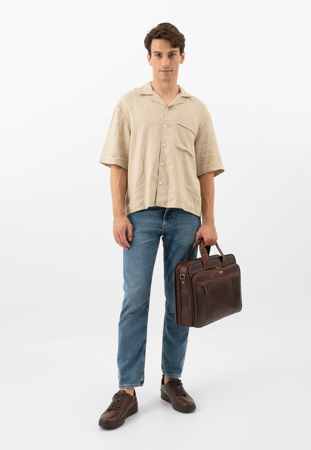 Model w beżowej koszuli i jeansach prezentuje ciemnobrązowe trampki męskie Wojas model 10149-52 z ozdobnym zamkiem, trzymając brązową teczkę biznesową.