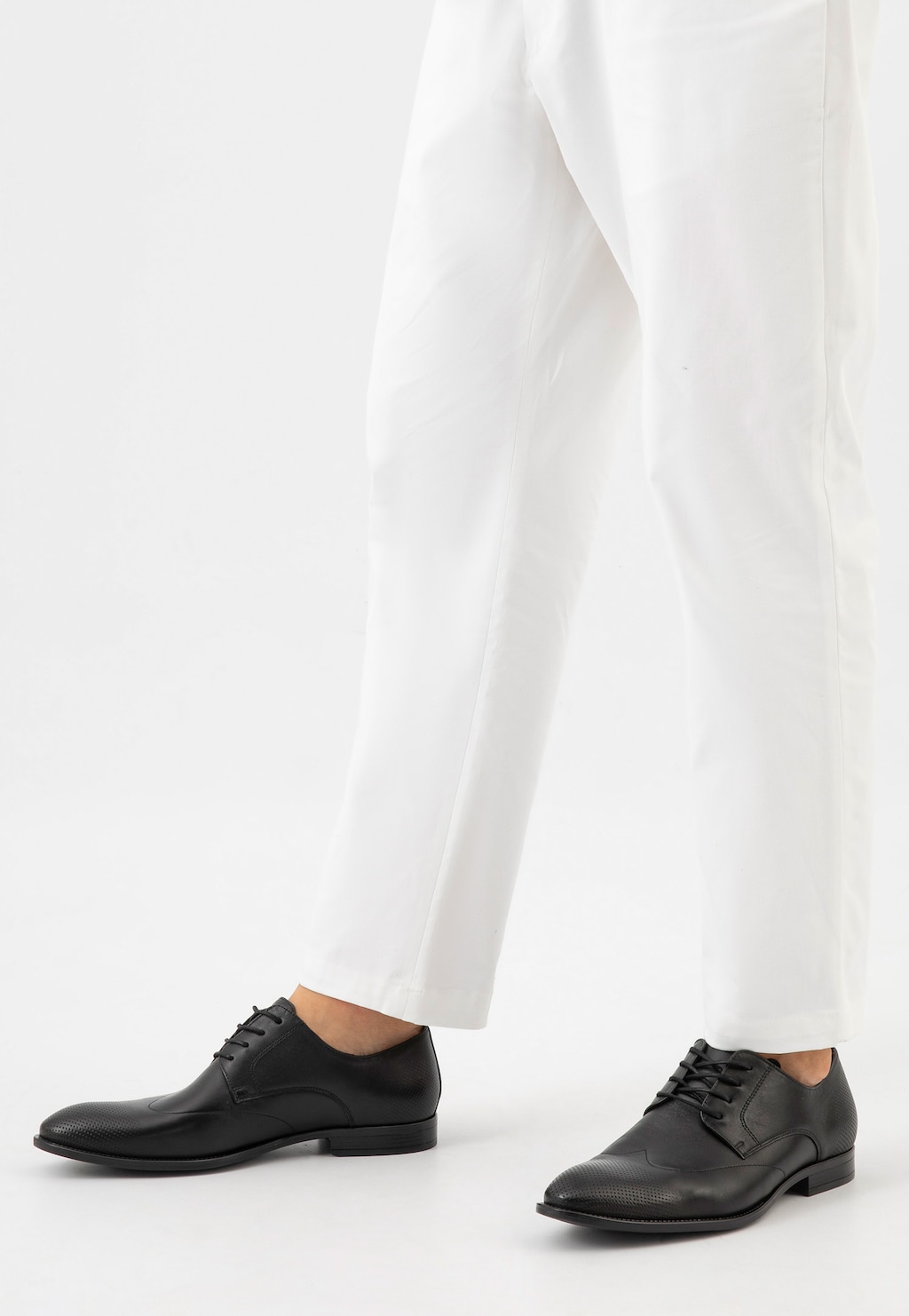 Czarne eleganckie buty męskie Wojas model 10195-51 ze skóry licowej z perforacją na czubku, prezentowane na nogach w białych spodniach, widok z boku.