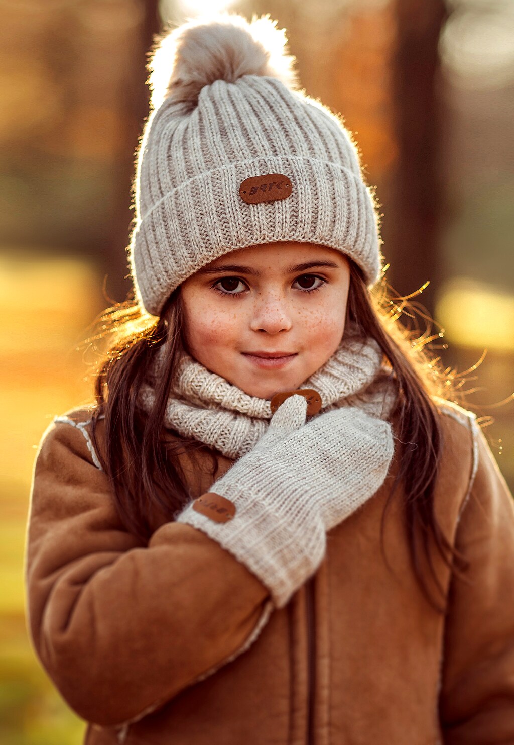 Dziecko w zimowym zestawie BARTEK 85601-14 składającym się z jasnoszarej czapki z pomponem, komina i rękawiczek, widoczne w ciepłym jesiennym świetle.