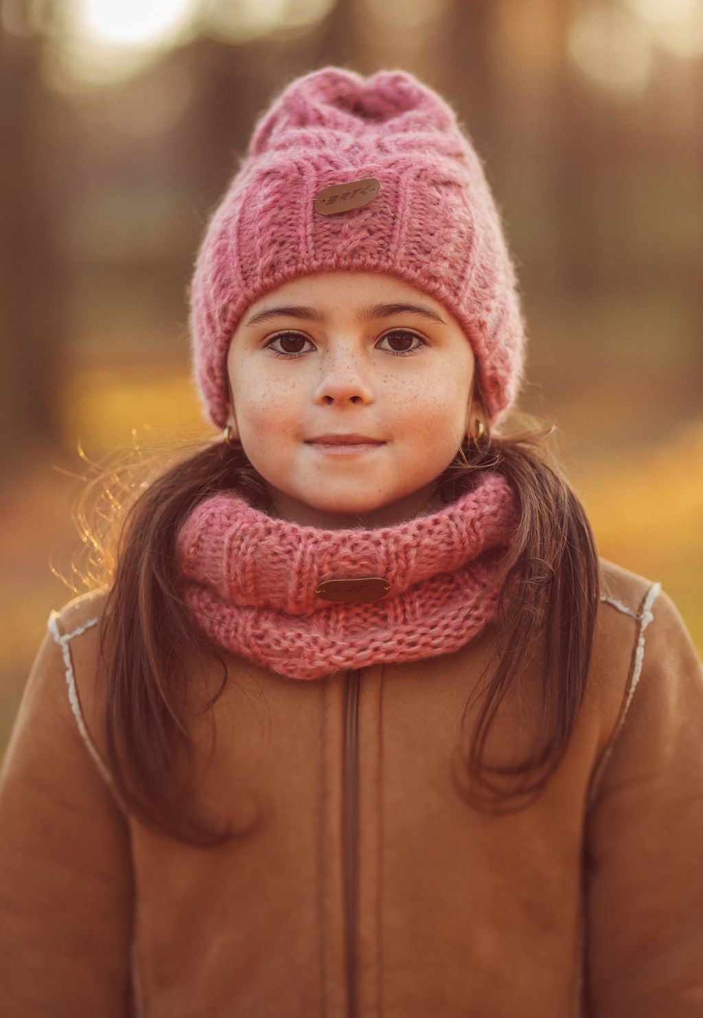Dziewczynka w różowym zestawie zimowym BARTEK 8560485 z wełny merino - czapka i komin w identycznym odcieniu. Dziecko ubrane w brązowy płaszczyk, jesienne tło.