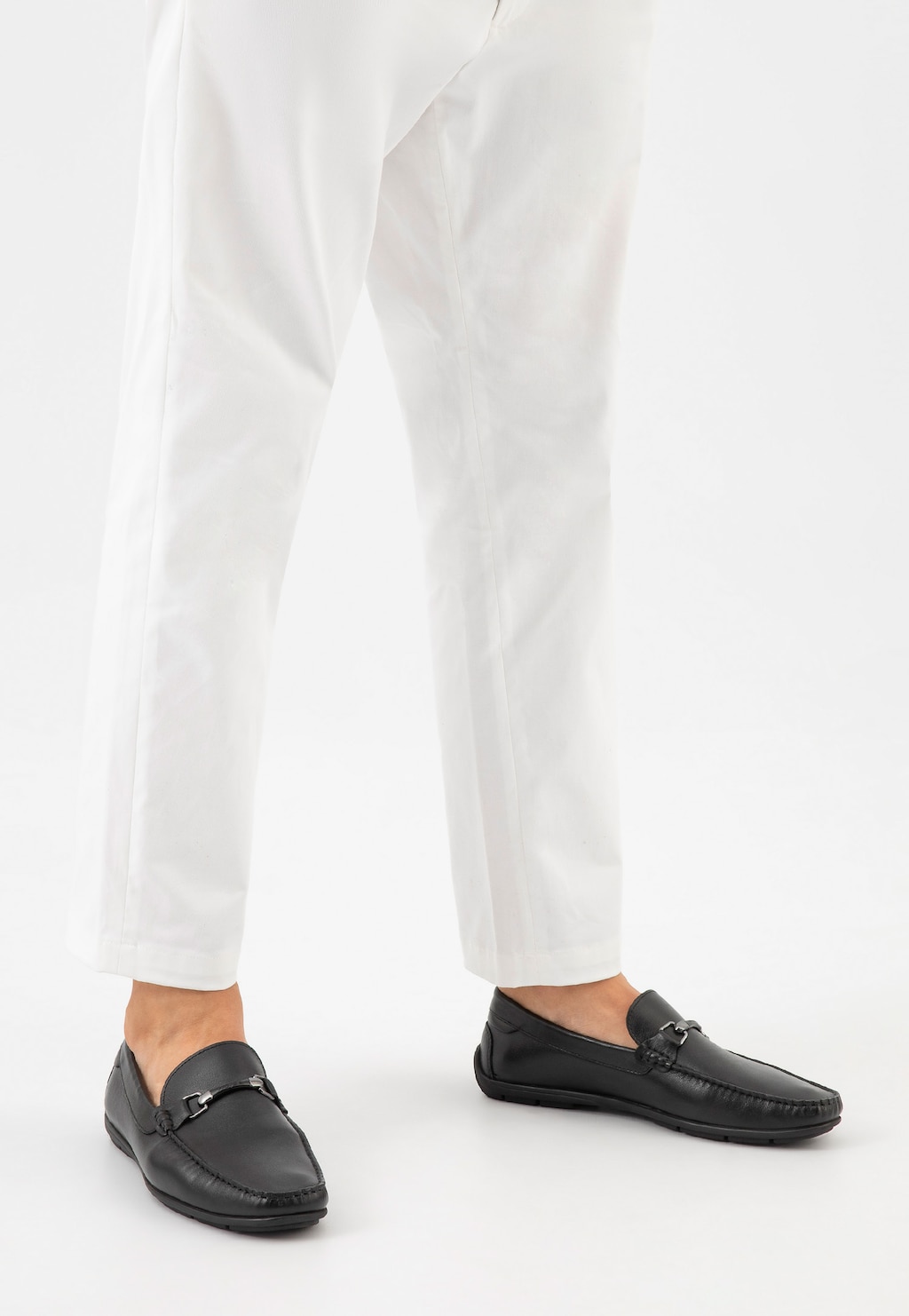 Czarne skórzane mokasyny męskie Wojas model 10200-51 z metalową ozdobą, noszone do białych spodni, widok z perspektywy frontalnej na stopach modela.