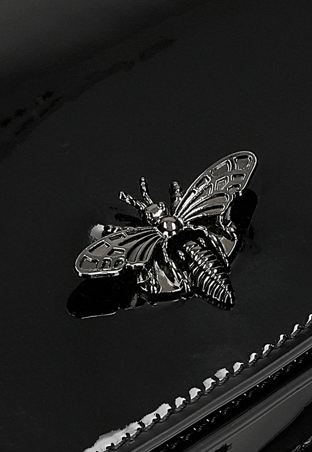 Metalowa ozdoba w kształcie owada (pszczoły lub motyla) na czarnej lakierowanej torebce Wojas model 80320-31, widoczne eleganckie wykończenie brzegów torebki.