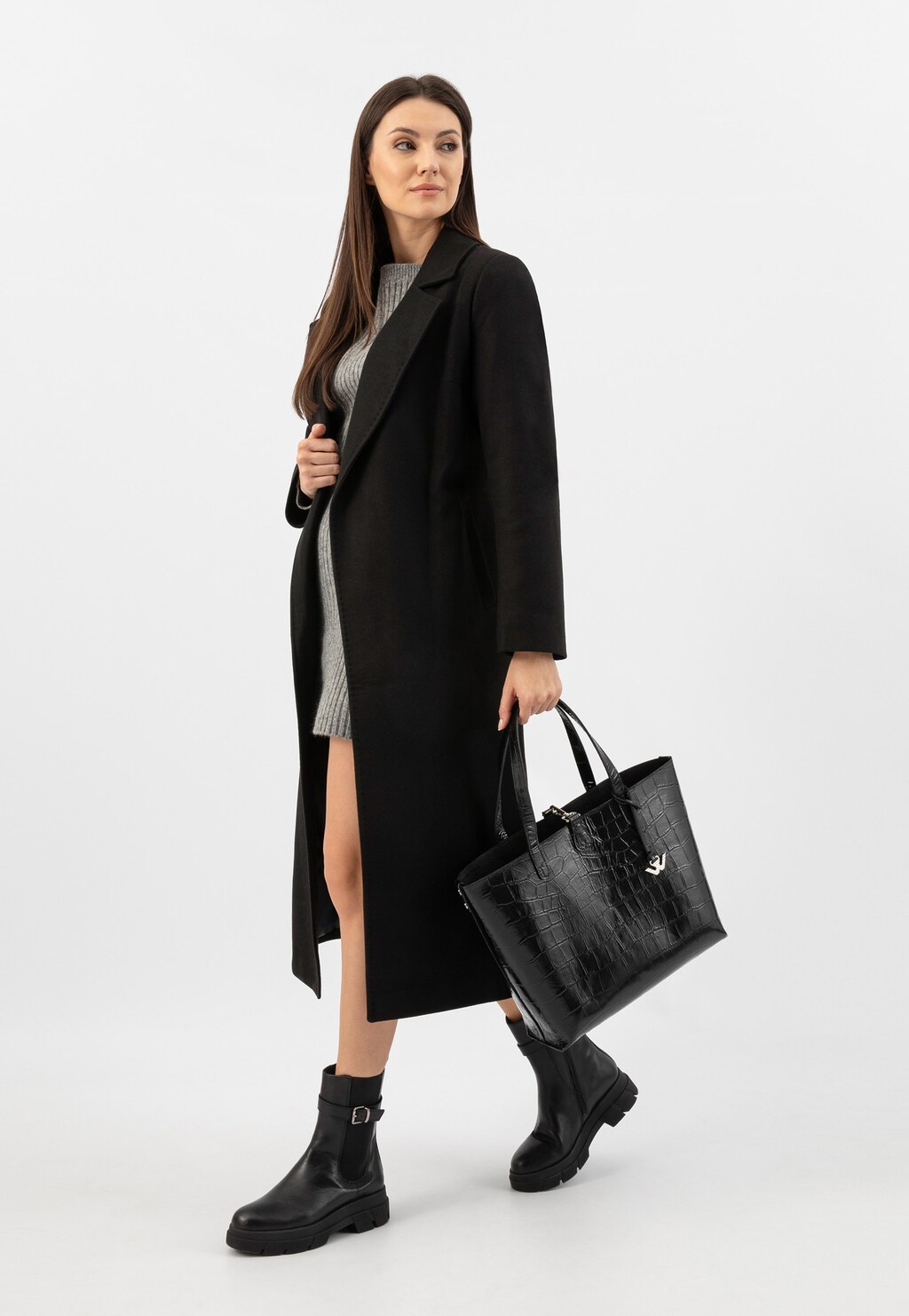Modelka w czarnym płaszczu i szarej sukience trzymająca dużą czarną torebkę Wojas 80250-81 z tłoczeniem w zwierzęcy print, uzupełniona czarnymi butami.