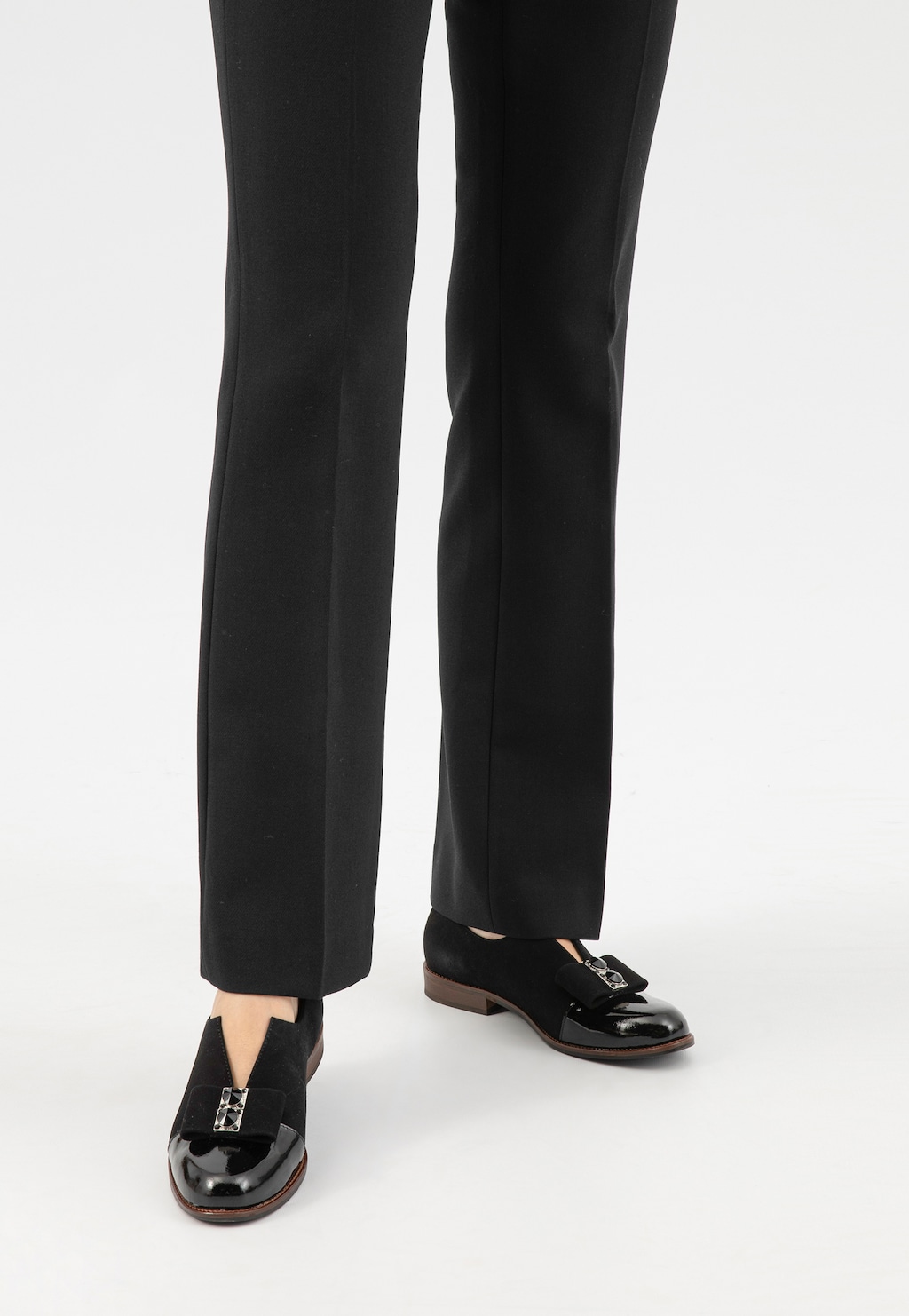 Czarne wsuwane półbuty damskie Wojas model 46302-71 z ozdobnym wycięciem i kokardą, widoczne na nogach w eleganckich czarnych spodniach, na brązowej podeszwie.