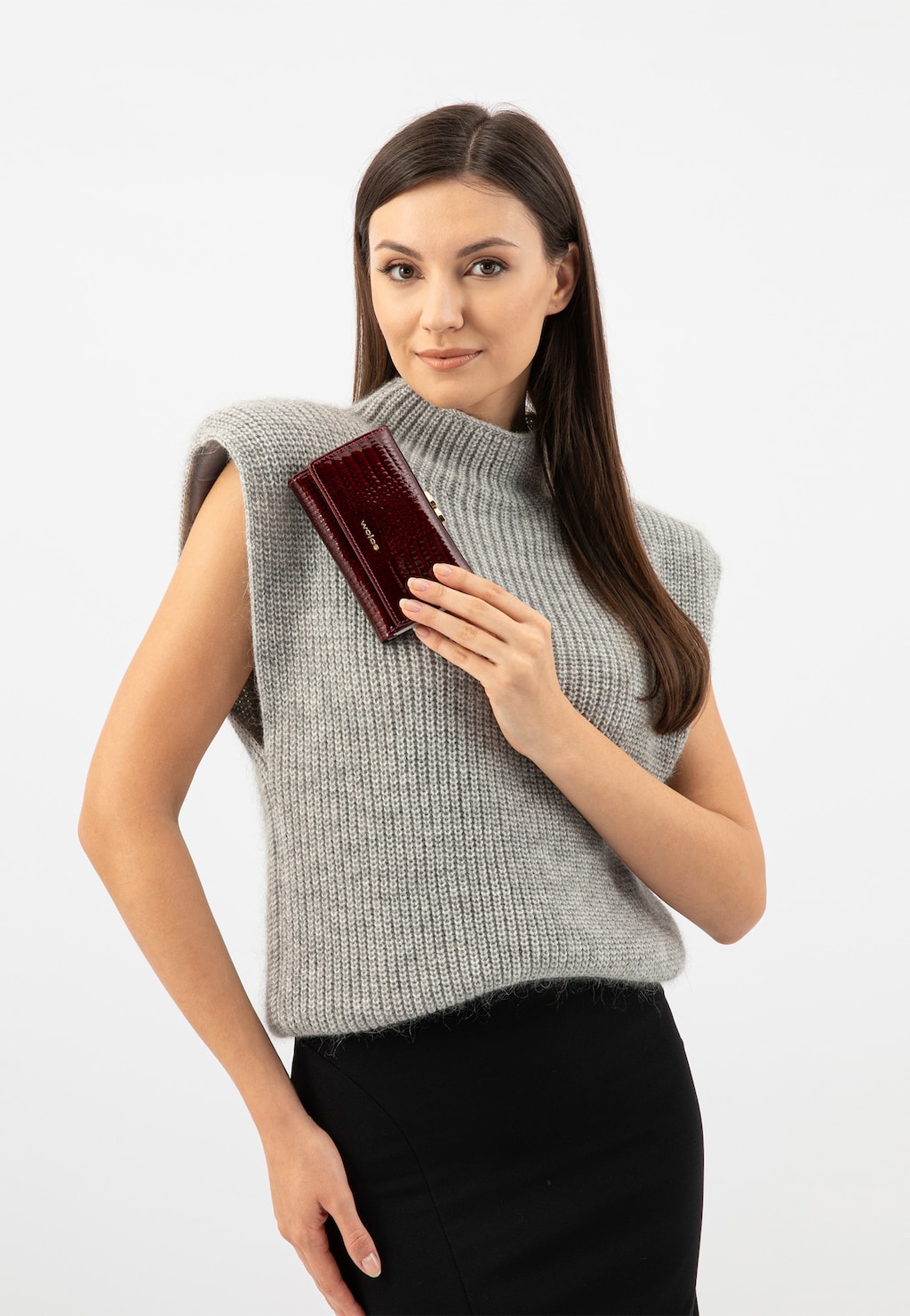 Elegancki czerwony portfel Wojas model 91085-35 trzymany przez modelkę w szarym swetrze bez rękawów. Lakierowana faktura portfela z wieloma przegrodami błyszczy w świetle.
