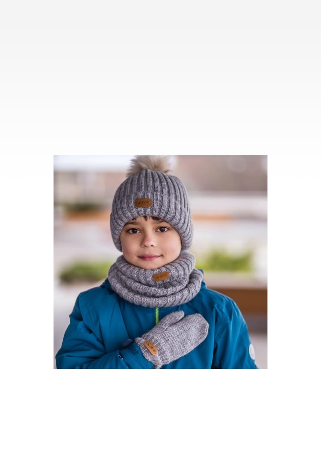 Popielaty zestaw zimowy BARTEK 85601-10 składający się z czapki z pomponem, komina i rękawiczek, prezentowany na tle rozmytego wnętrza.