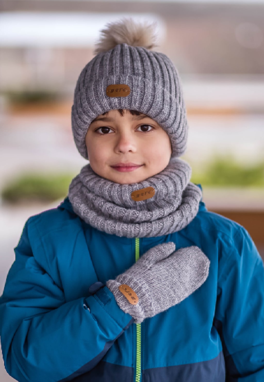Popielaty zestaw BARTEK 85601-10 z dzianinową czapką z pomponem, kominem i rękawiczkami, prezentowany na dziecku w niebieskiej kurtce, z widocznymi logo marki na każdej części.