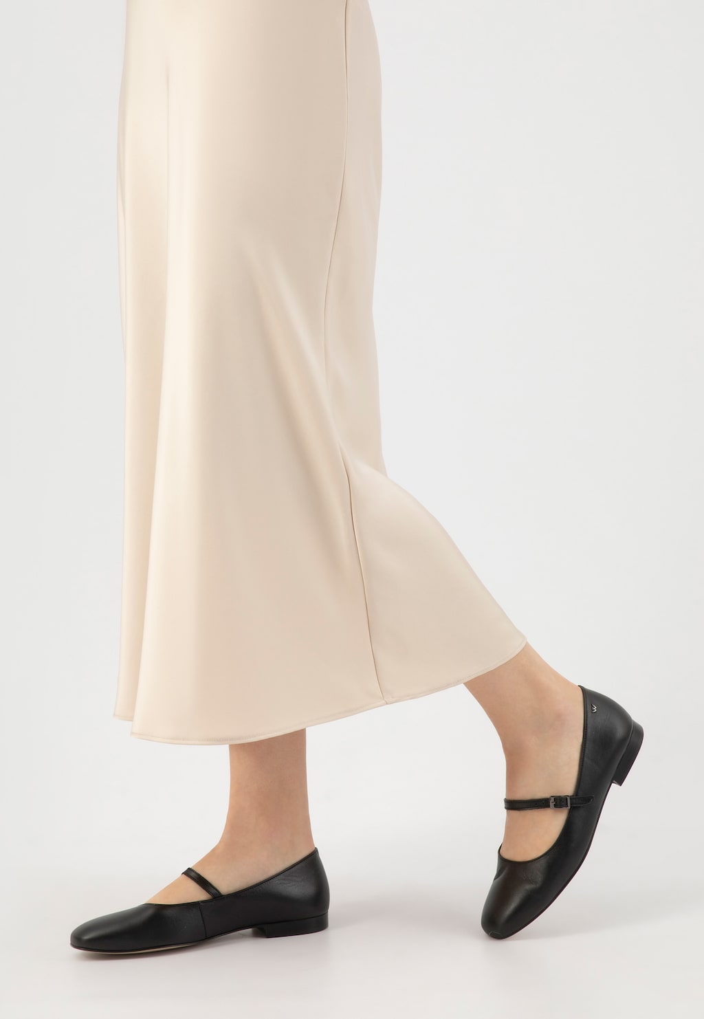 Czarne skórzane baleriny Wojas 44029-51 z eleganckim paskiem na podbiciu, noszone do kremowej spódnicy, pokazane z boku na stopach modelki.