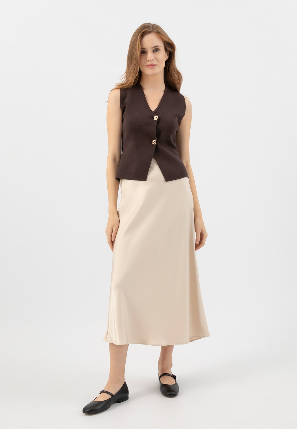 Modelka w brązowej kamizelce i jasnej spódnicy midi, prezentująca czarne skórzane baleriny damskie Wojas z paskiem na podbiciu, model 44029-51.