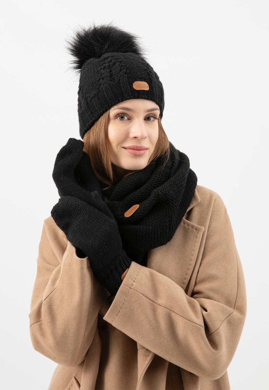 Czarny zimowy zestaw Relaks R96018-11 - czapka z pomponem, komin i rękawiczki, prezentowane na modelce w beżowym płaszczu. Zestaw zdobią eleganckie brązowe metki.