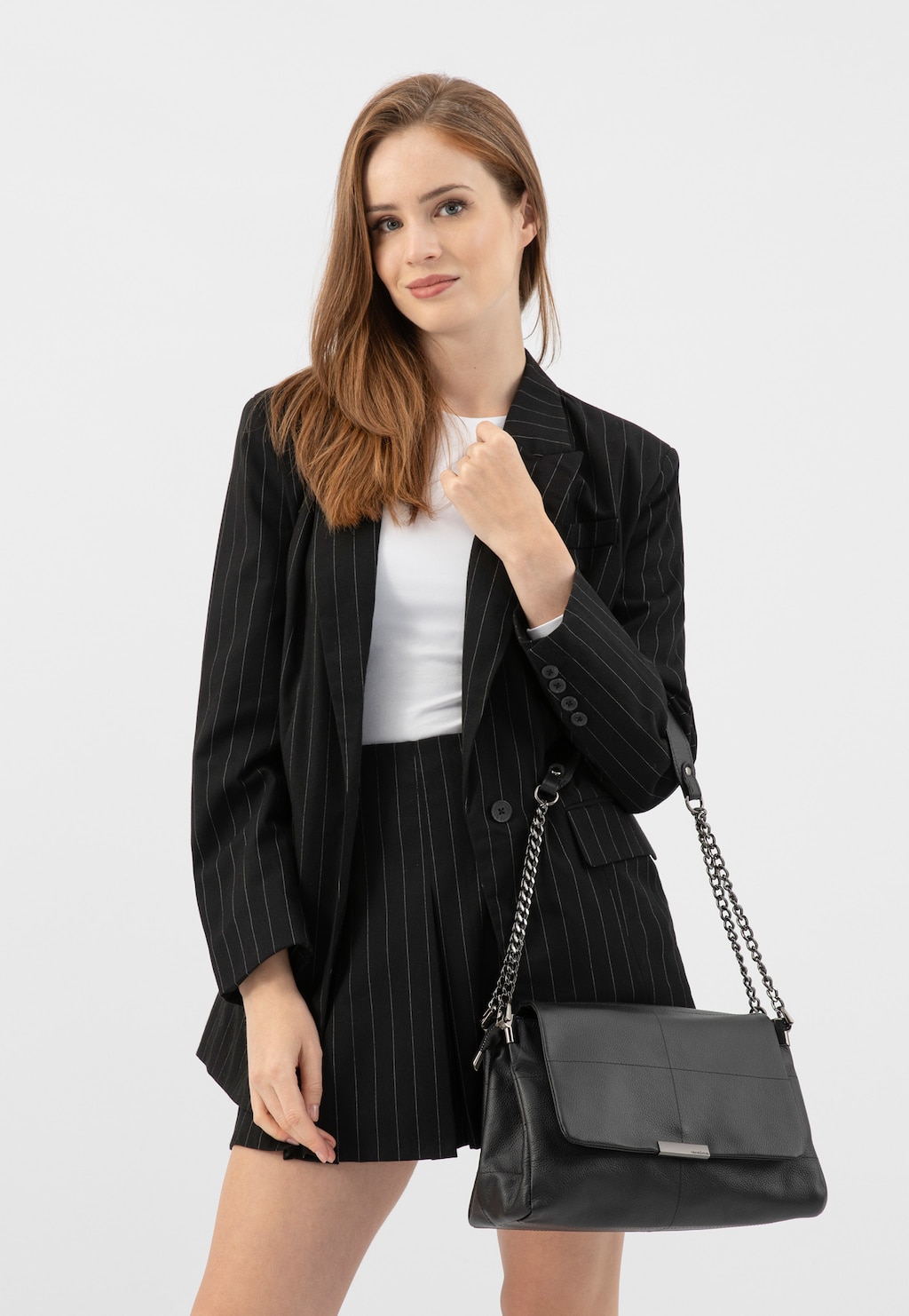 Elegancki czarny garnitur w paski z białym topem, uzupełniony czarną skórzaną torebką Wojas model 80416-51 z charakterystycznym łańcuszkiem na pasku.