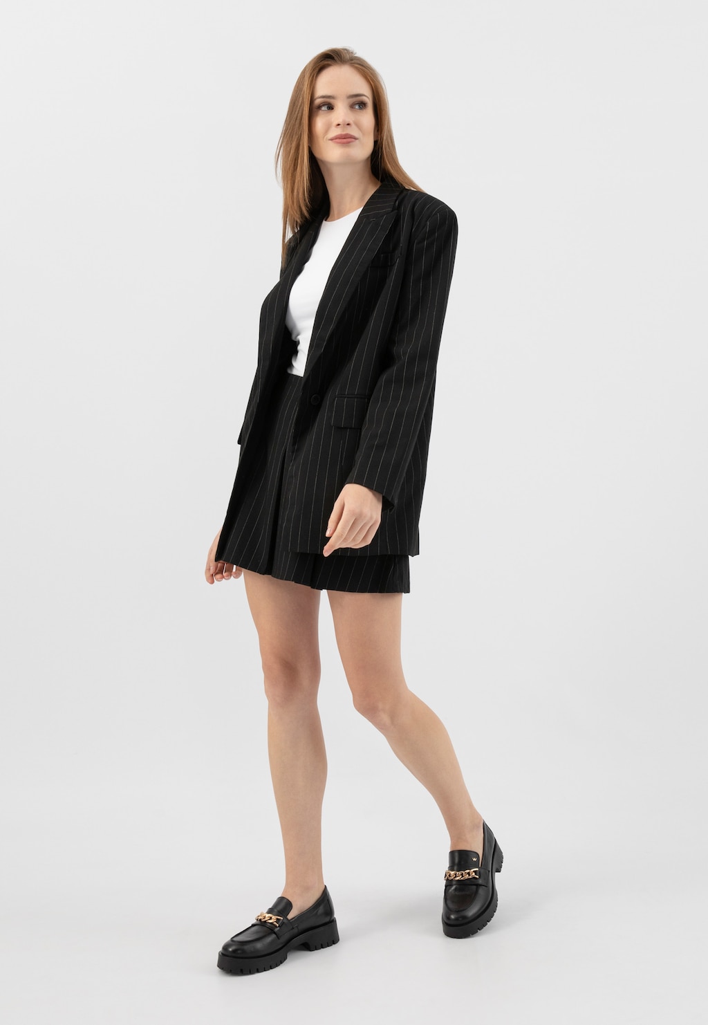 Modelka w czarnym garniturze w paski i białym topie, prezentująca czarne mokasyny damskie Wojas 46323-51 ze skóry licowej ze złotą ozdobą na przodzie buta.