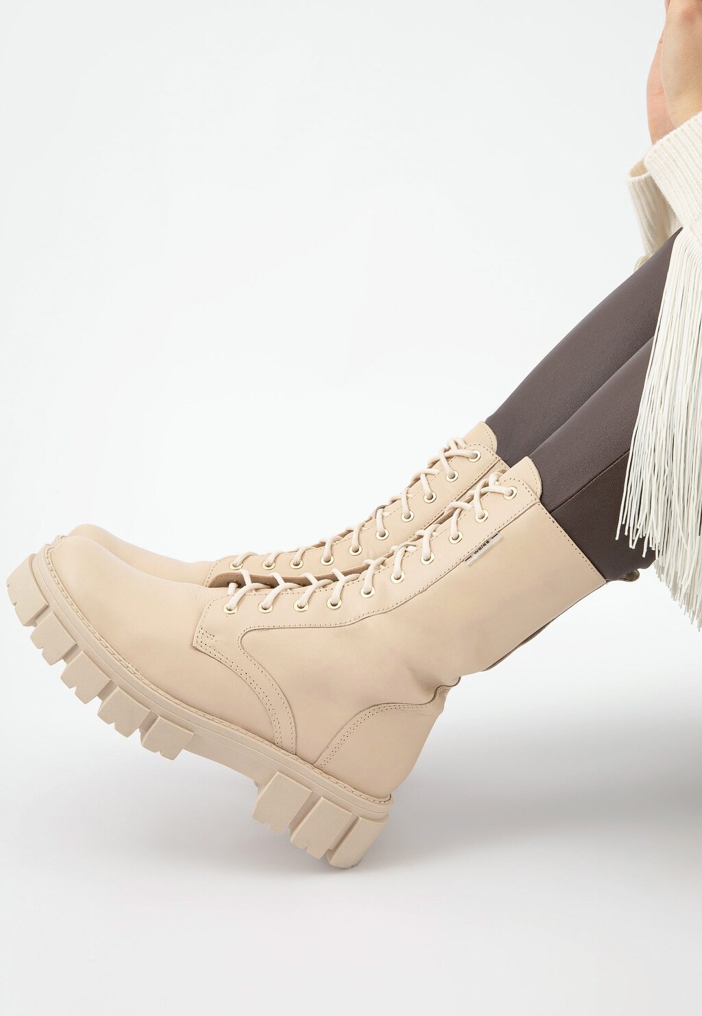 Wysokie beżowe trzewiki damskie Wojas model 64020-54 typu bikery z grubą podeszwą, sznurowane, prezentowane na nodze z ciemnymi rajstopami i białą frędzlowaną górą.