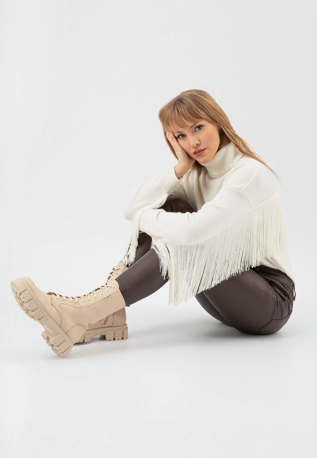 Modelka w białym swetrze z frędzlami prezentuje wysokie beżowe trzewiki damskie typu bikery Wojas model 64020-54 z masywną podeszwą i sznurowaniem.