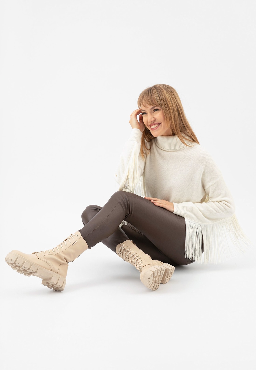 Kobieta w białym swetrze i brązowych legginsach prezentuje wysokie beżowe trzewiki damskie typu bikery Wojas model 64020-54, uśmiechając się i siedząc na podłodze.