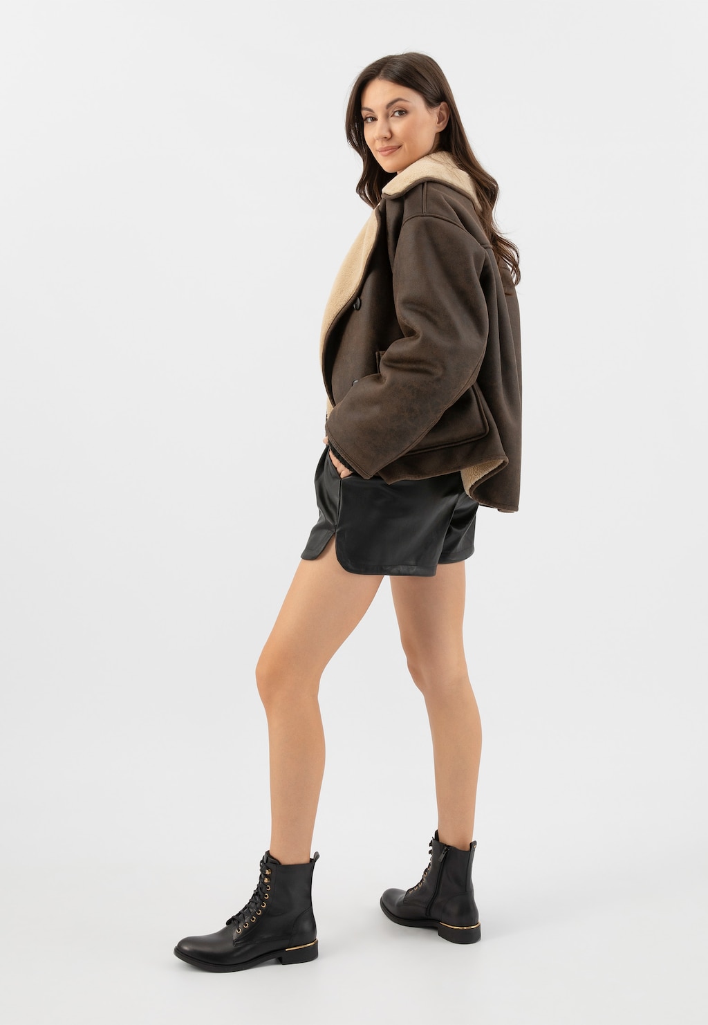 Modelka w brązowej kurtce kożuchowej i czarnych szortach, prezentująca czarne skórzane trzewiki Wojas model 64127-51 ze złotymi elementami, widoczne na stopach.