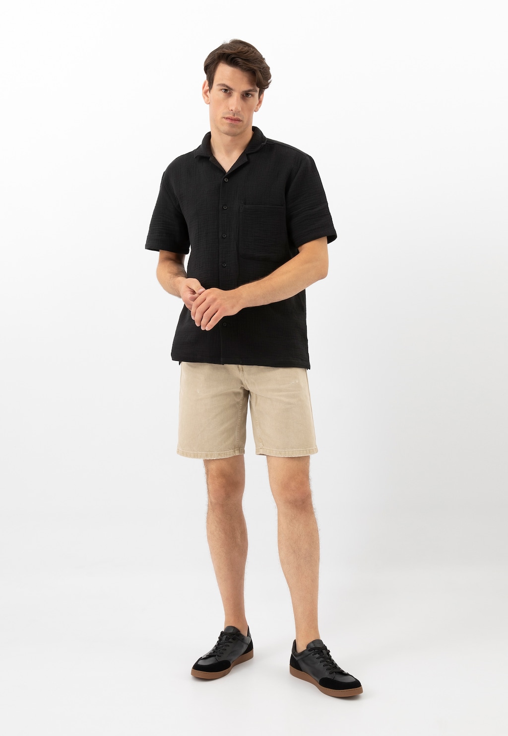 Model prezentuje czarne trampki Wojas 10217-51 z łączonych skór na brązowej podeszwie, zestawione z czarną koszulą i beżowymi szortami.