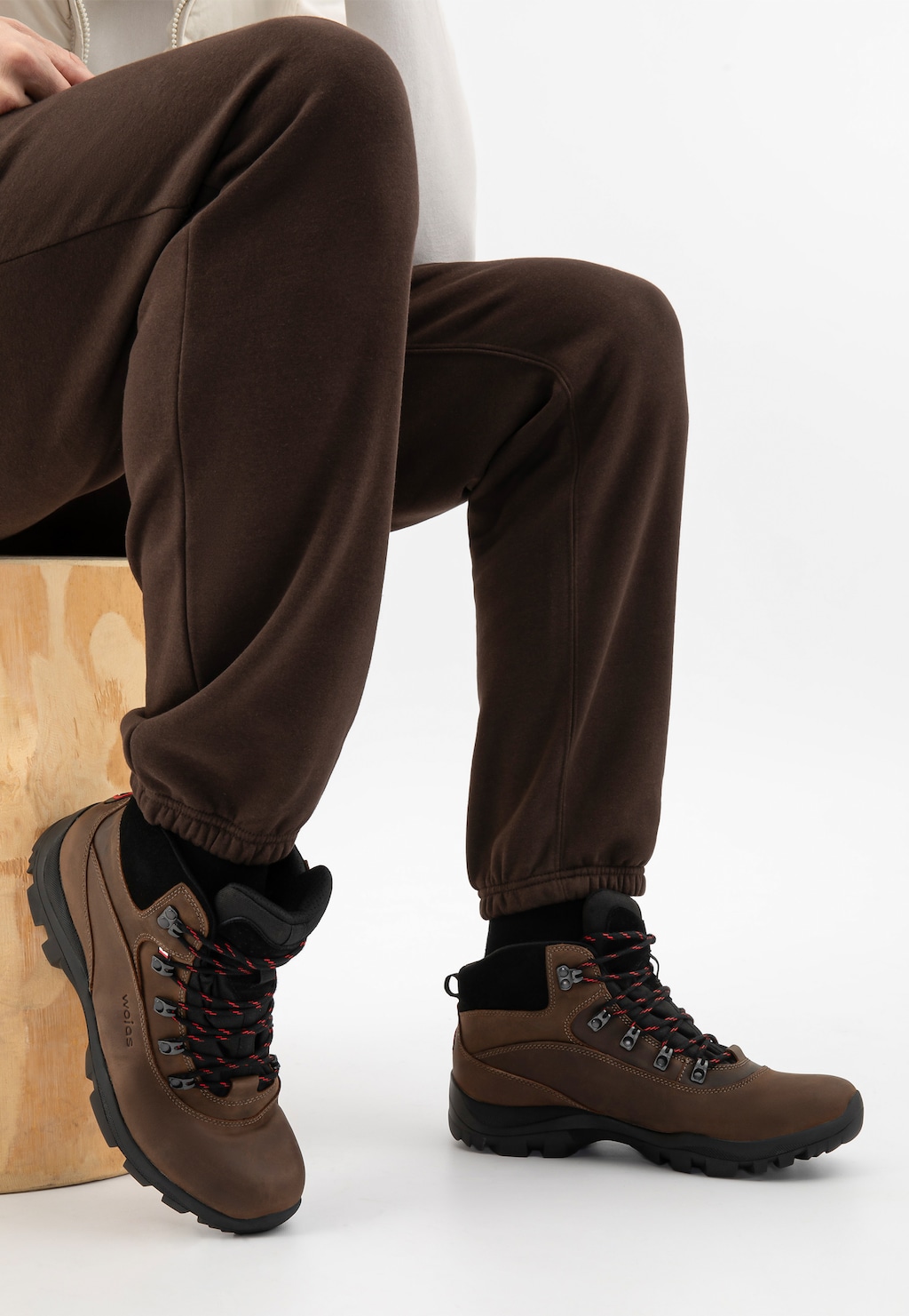 Brązowe męskie trzewiki trekkingowe Wojas model 24027-92 na nogach mężczyzny siedzącego na drewnianym stołku, ubrane do brązowych spodni dresowych.