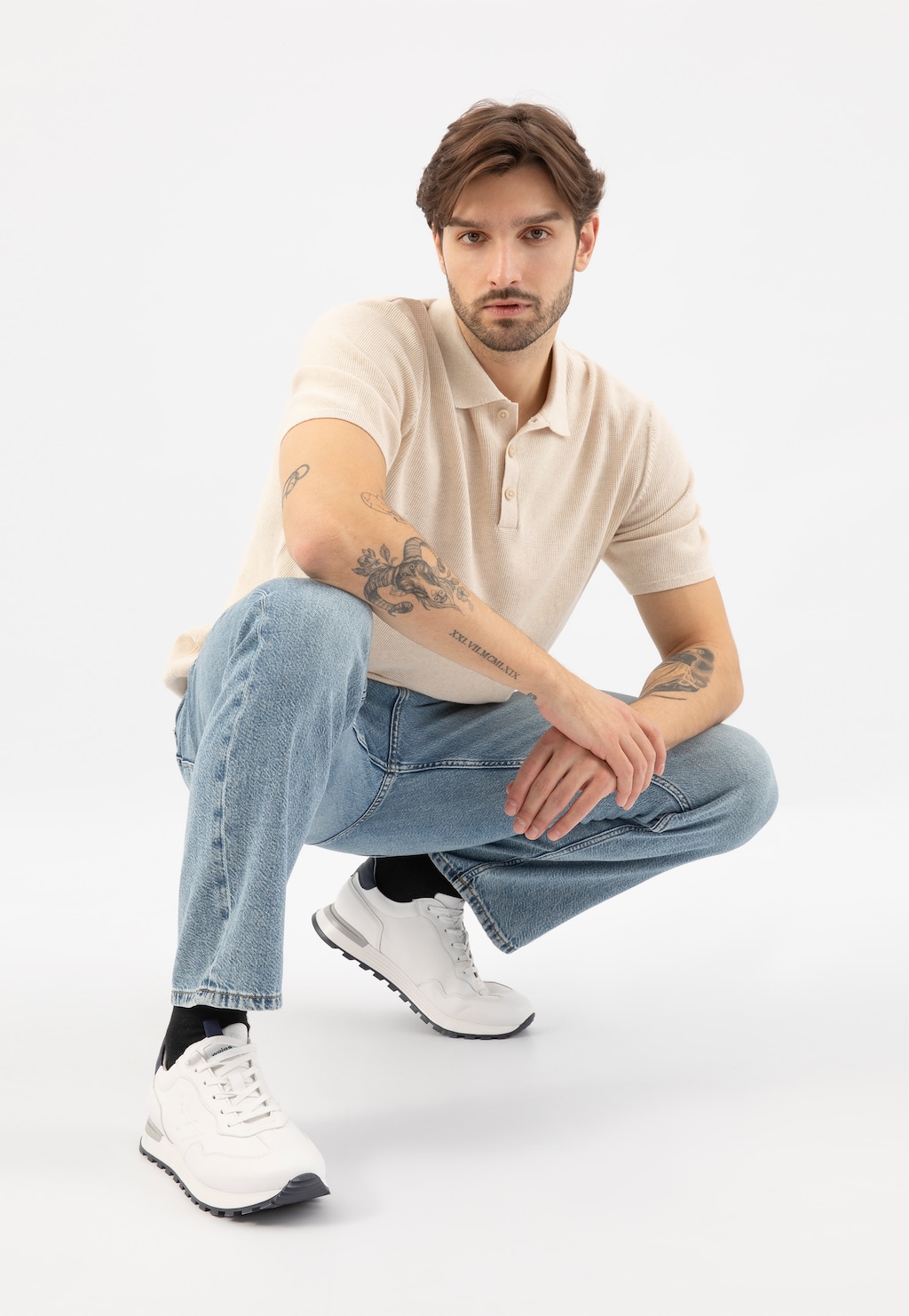 Osoba w kucki w kremowej koszulce polo i jeansach, prezentująca białe sneakersy Wojas model 10201-50 na kontrastowej czarnej podeszwie, idealne do casualowych stylizacji.