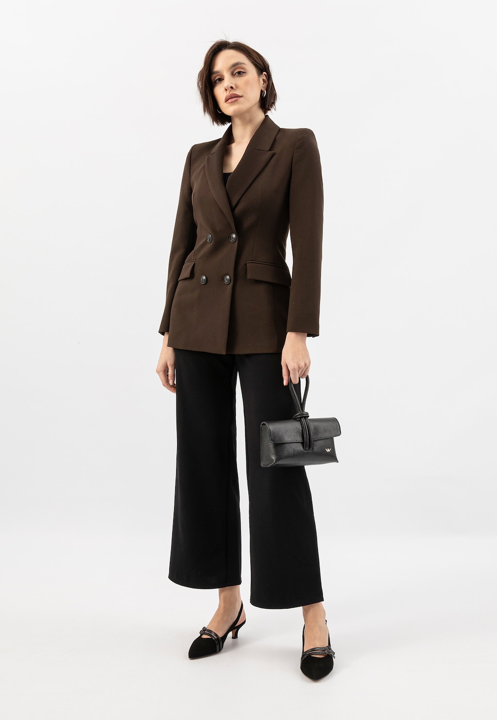 Modelka w brązowej marynarce i czarnych spodniach prezentuje elegancką czarną torebkę Wojas model 80430-51, trzymając ją w dłoni. Stylowy biznesowy outfit uzupełniają czarne buty.