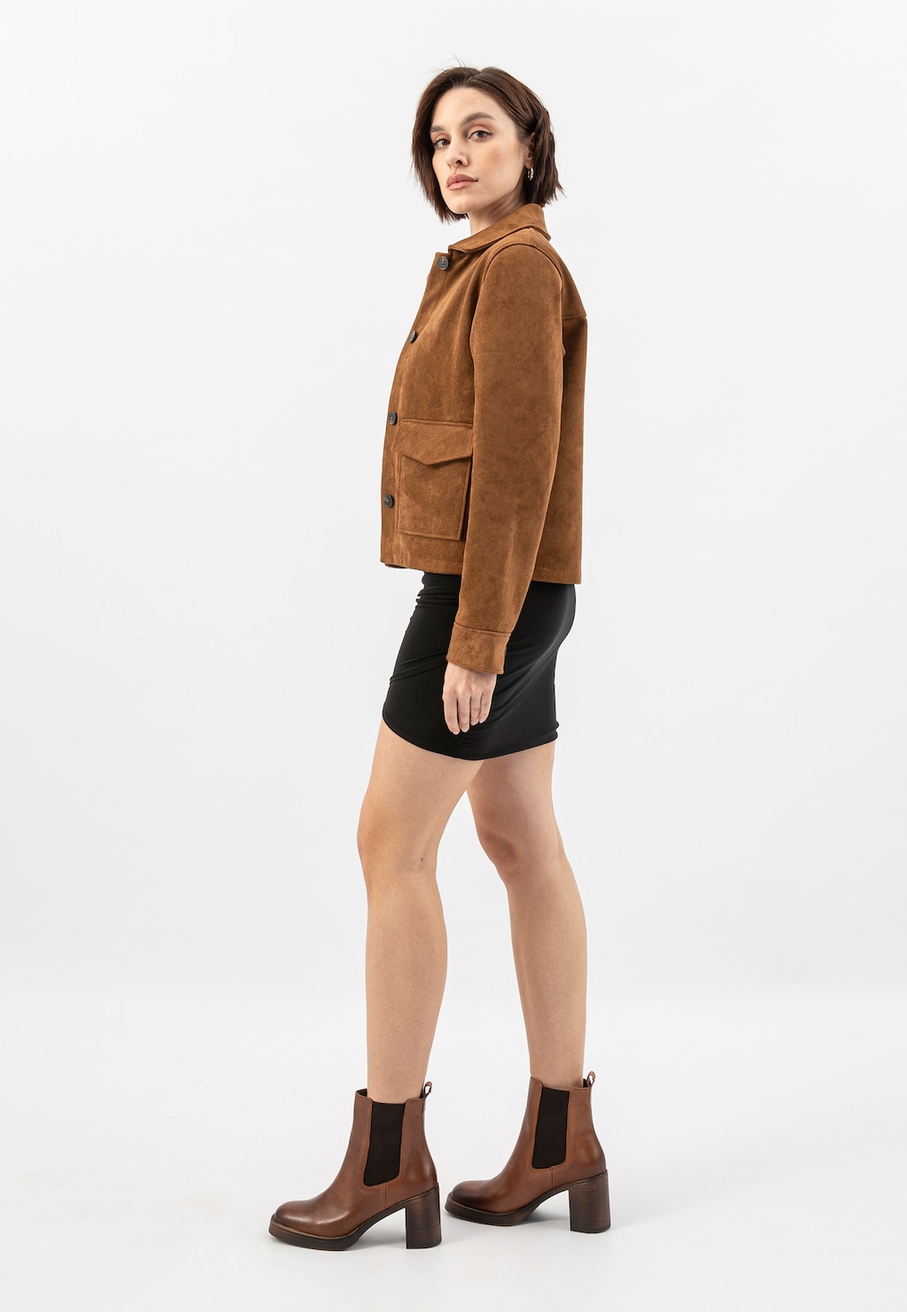 Modelka w brązowej zamszowej kurtce i czarnej spódnicy, prezentująca brązowe skórzane sztyblety Wojas model 55260-52 na obcasie, widoczne z boku na nogach.