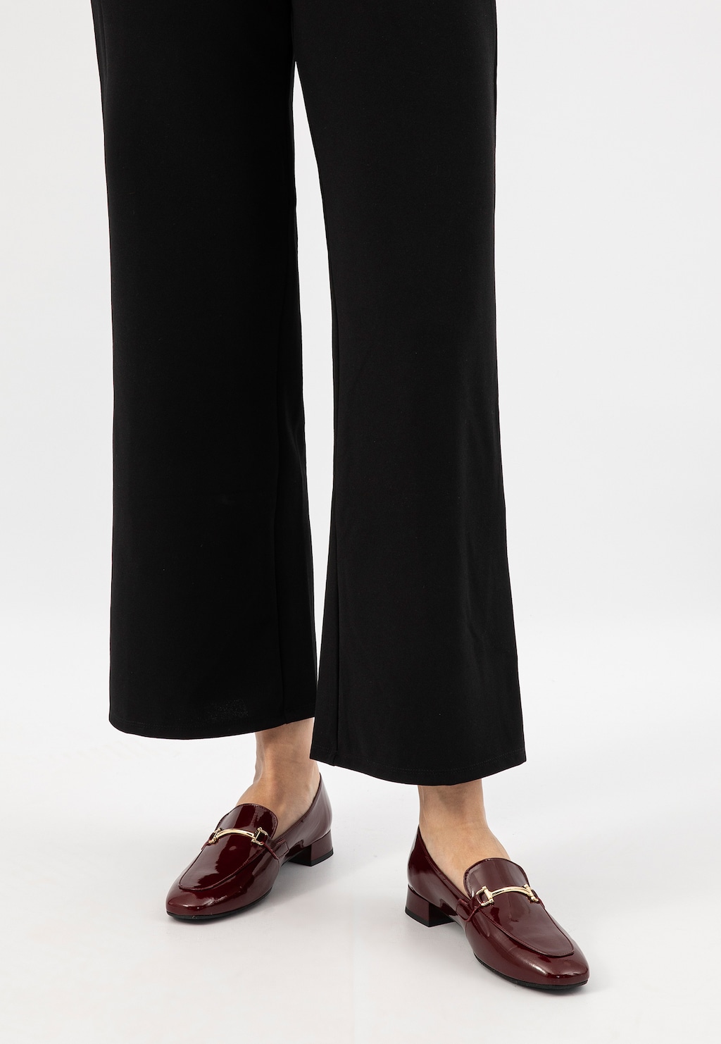 Bordowe lakierowane mokasyny damskie Wojas 46348-35 na niskim obcasie, noszone do eleganckich czarnych spodni, widok od dołu na stopy modelki.