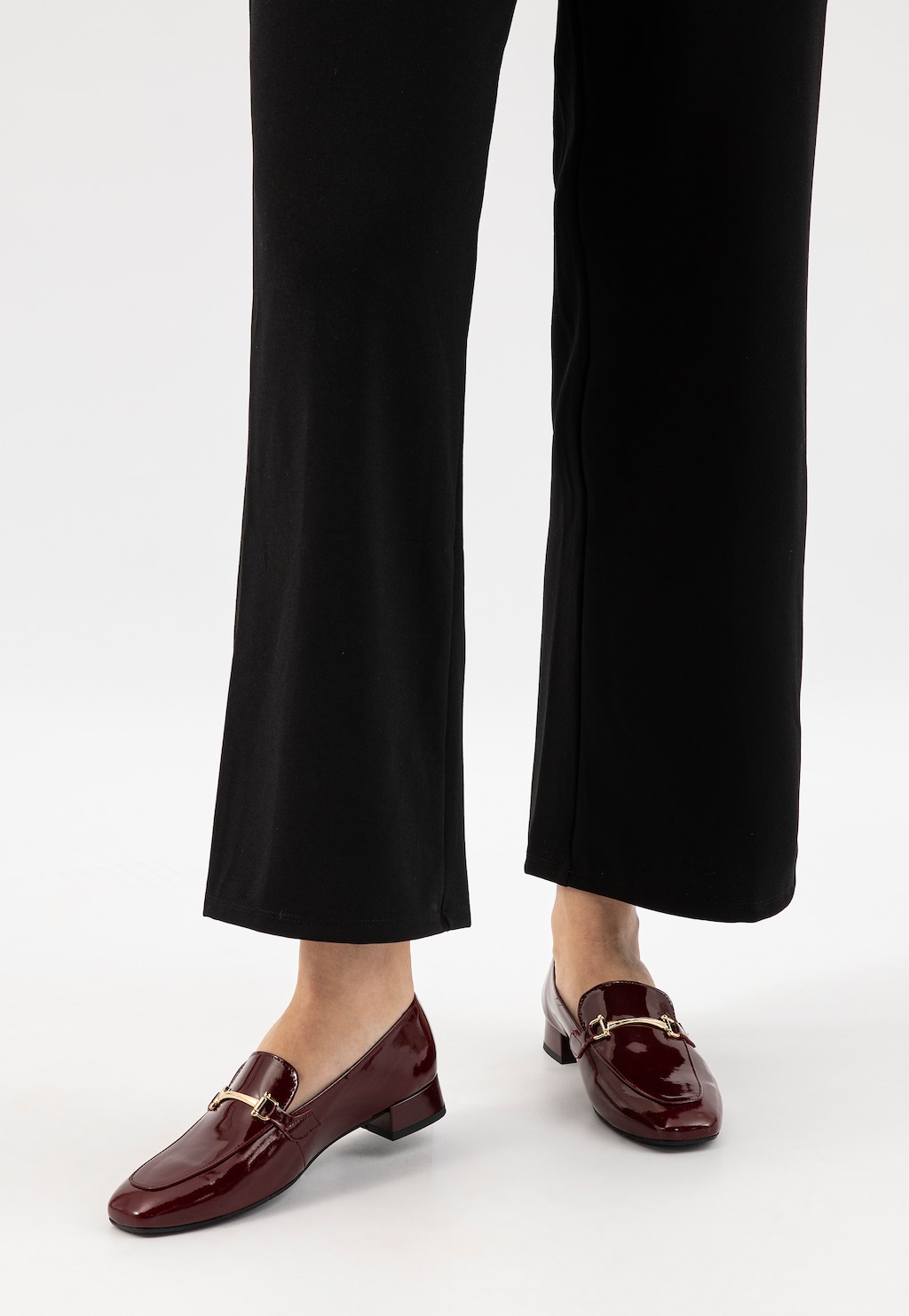 Bordowe lakierowane mokasyny damskie Wojas model 46348-35 na niskim obcasie, widoczne na nogach w czarnych spodniach, ze złotą klamrą zdobiącą przód buta.