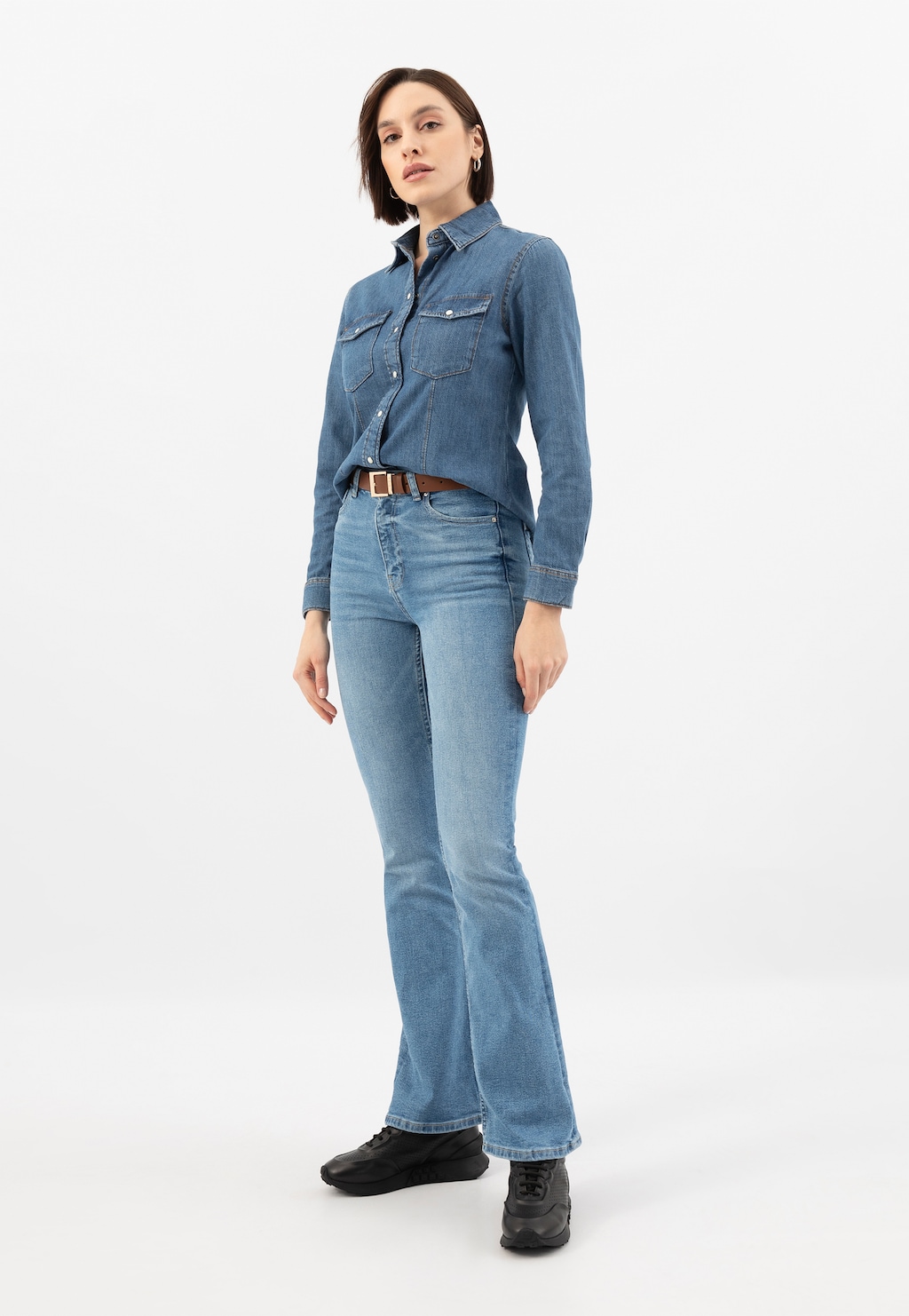 Modelka w jeansowym stroju prezentuje czarne skórzane sneakersy damskie Wojas model 46306-51. Buty widoczne z przodu, stanowią uzupełnienie casualowej stylizacji z dżinsową koszulą i spodniami.