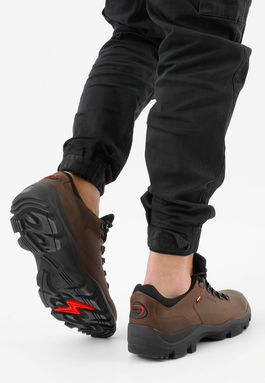 Brązowe męskie półbuty trekkingowe Wojas model 9377-92 na nogach osoby w czarnych spodniach, widoczna wytrzymała podeszwa z czerwonym elementem, idealne do aktywności outdoorowych.