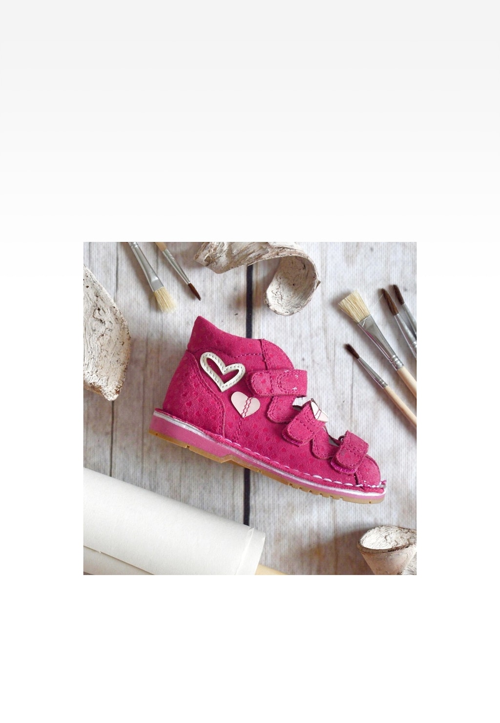 Różowe sandały zabudowane BARTEK 8550065 dla dziewcząt z ozdobnymi rzepami i aplikacją w kształcie serca, sfotografowane z boku wśród pędzli malarskich na drewnianej powierzchni.