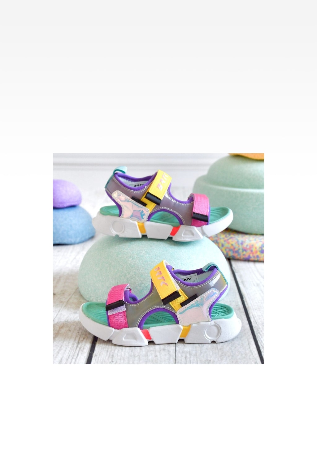 Kolorowe sandały dziecięce Bartek 16043202 dla dziewcząt, z zapięciem na rzepy w odcieniach fioletu, różu i żółci, na białej podeszwie, ustawione na pastelowych dekoracjach.
