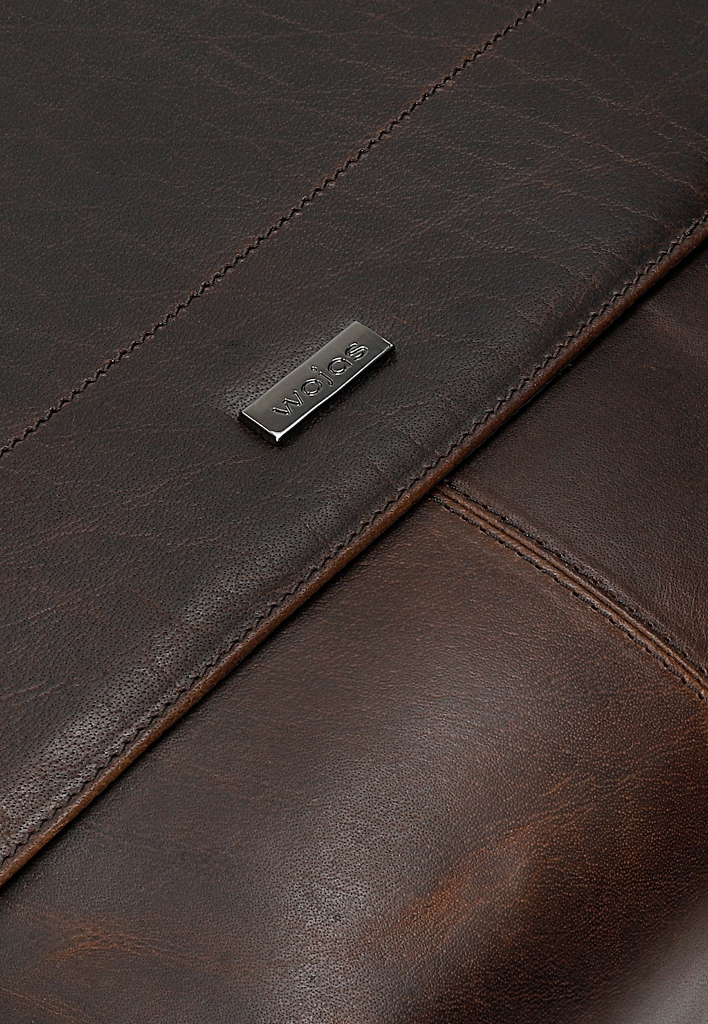 Zbliżenie na duży, brązowy fragment torby męskiej Wojas model 90025-42 z woskowanej skóry typu pull up, widoczny metalowy znaczek marki i staranne przeszycia.