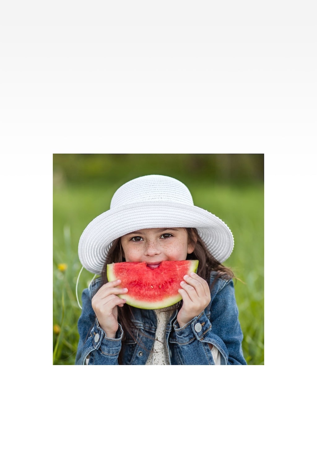 Dziewczynka w białym plecionym kapeluszu Bartek 85607-19 jedząca arbuza na zielonej łące, ubrana w jeansową kurtkę. Letnia stylizacja z kapeluszem przeciwsłonecznym.