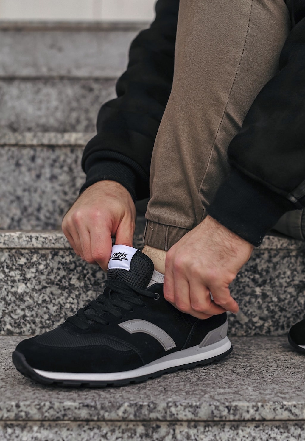 Mężczyzna zakładający czarne sportowe sneakersy RELAKS R34202-89 z szarymi wstawkami, siedzący na kamiennych schodach.