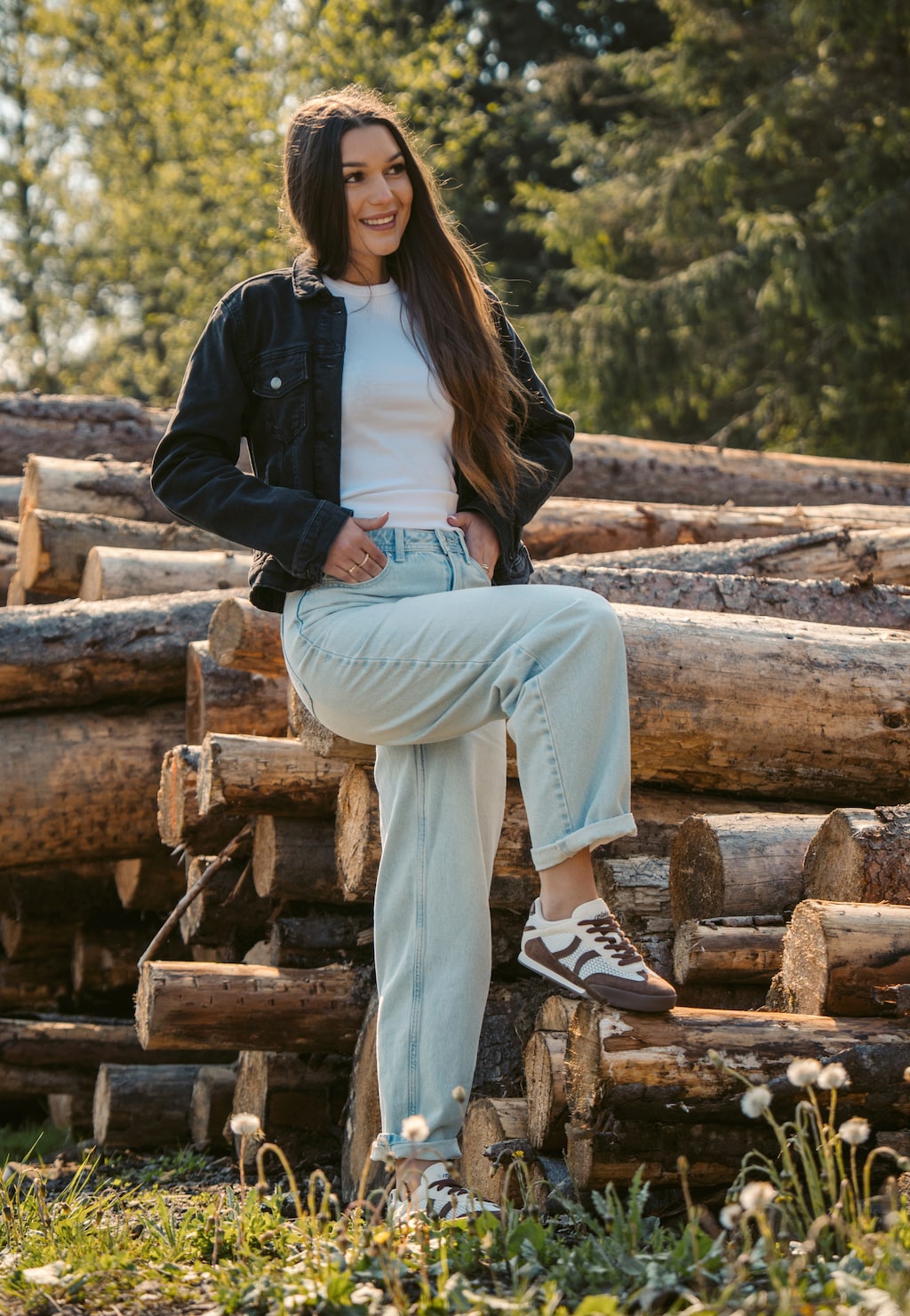 Kobieta siedząca na kłodach drewna w beżowo-brązowych sneakersach damskich RELAKS R46185-82 w stylu retro z ażurową cholewką, jeansy i kurtka denim.