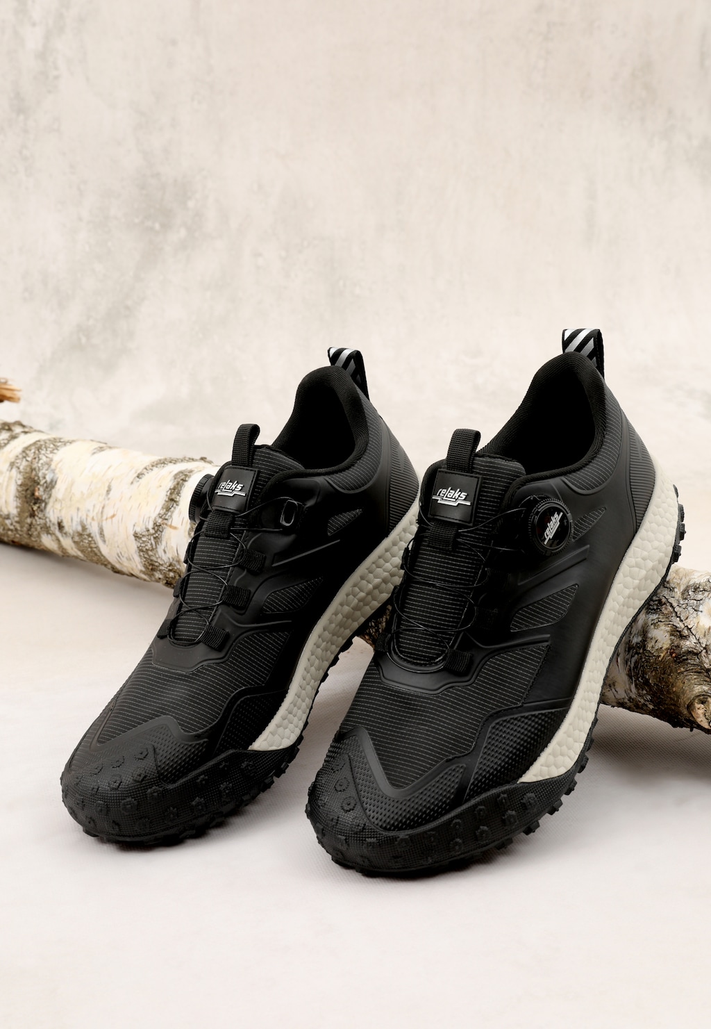 Czarne sneakersy RELAKS R10125-11 ze sznurowaniem na pokrętło, białą podeszwą i bieżnikiem terenowym, widok z boku na tle drewnianych gałęzi.