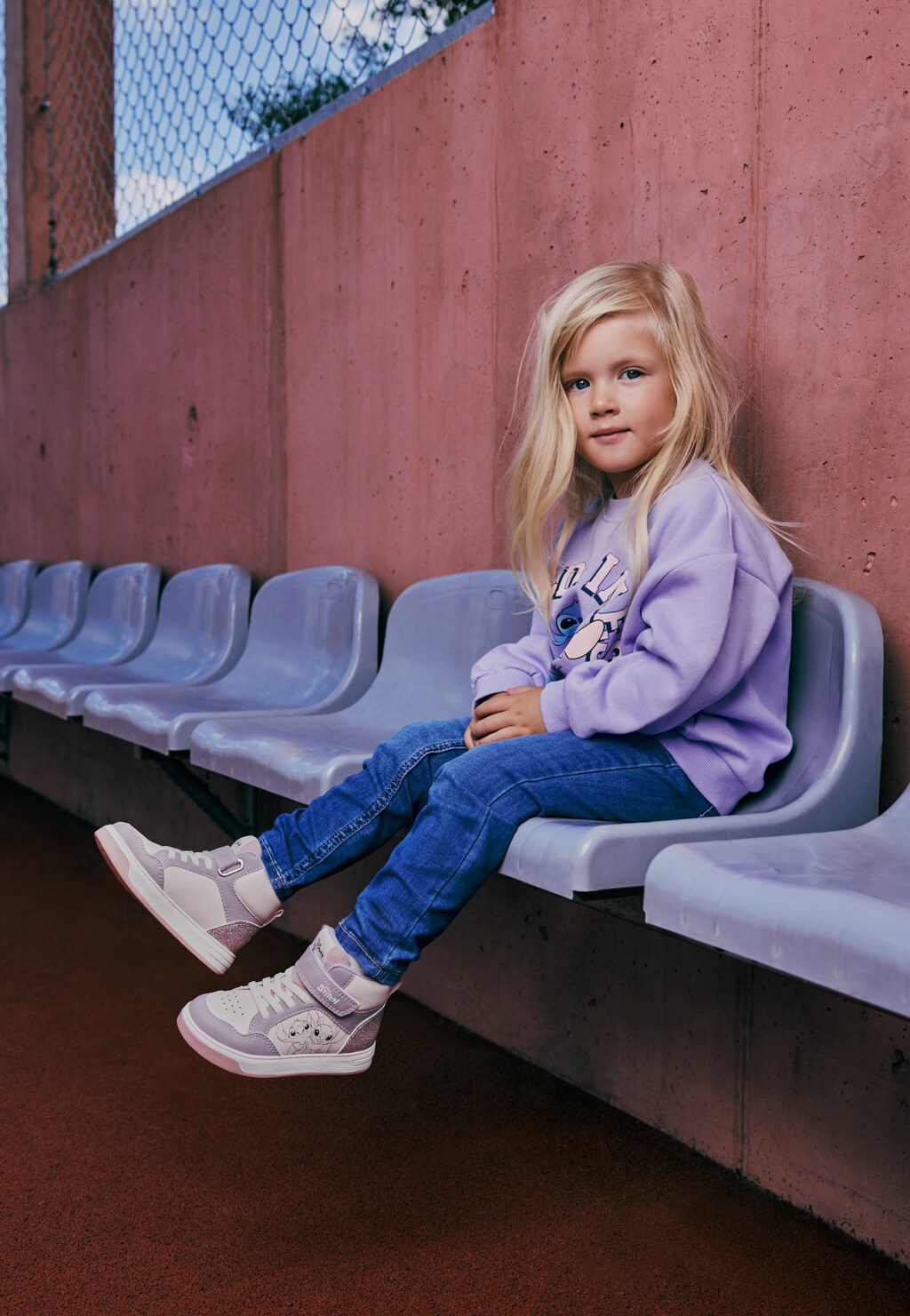 Dziewczynka siedząca na trybunach w fioletowych wysokich trampkach Stitch Bartek 88019-16, bluzie ze Stitchem i jeansach.
