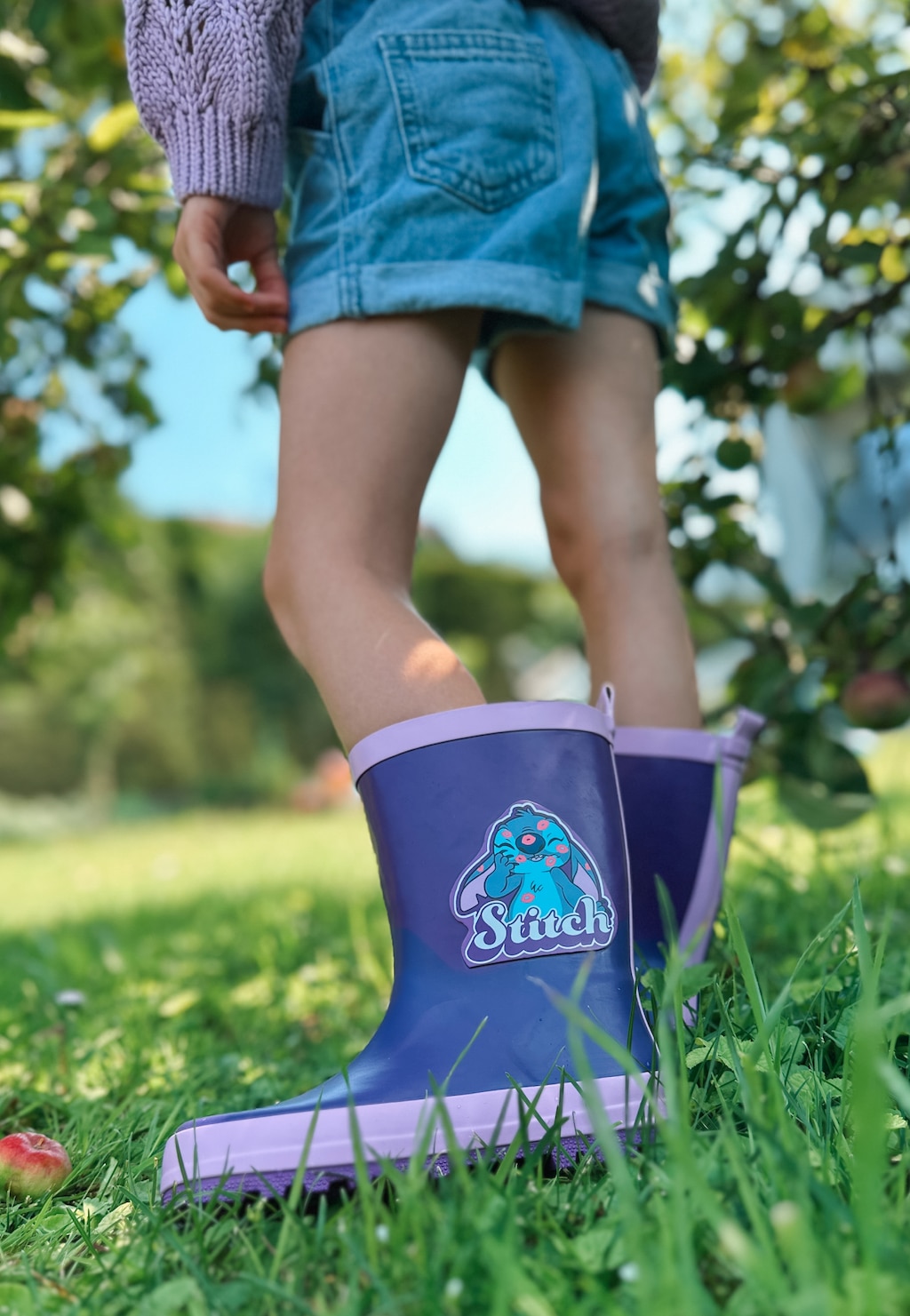 Fioletowe kalosze dziewczęce Bartek Stitch 86700-16 z nadrukiem Lilo & Stitch na nogach dziecka stojącego na trawie w ogrodzie.