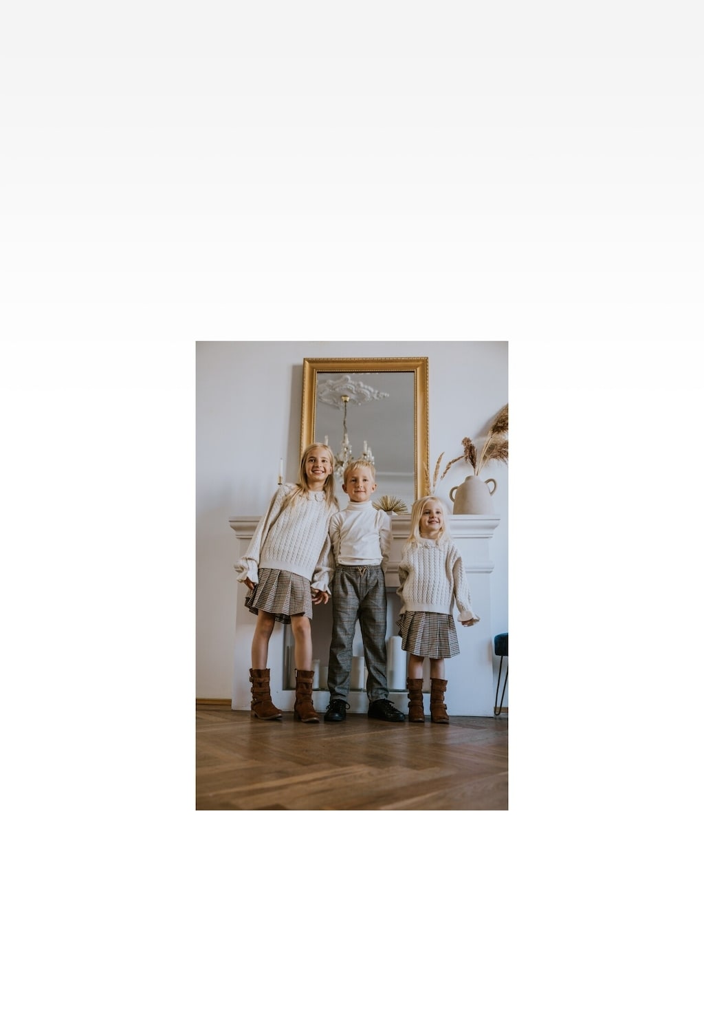 Troje dzieci w wełnianych swetrach i spódniczkach/spodniach w kratkę, noszących brązowe botki Wojas x Bartek 8440263, pozujących na tle lustra w złotej ramie i jasnej ściany.