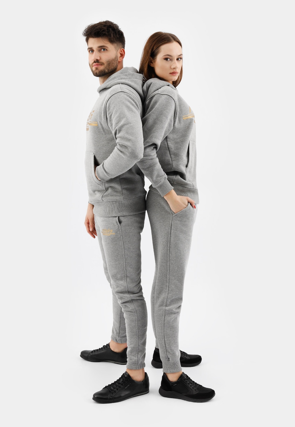 Popielate spodnie dresowe damskie model 98024-80 prezentowane na modelce, stojącej plecami do modela. Oboje w dopasowanych szarych dresach i butach marki Relaks.