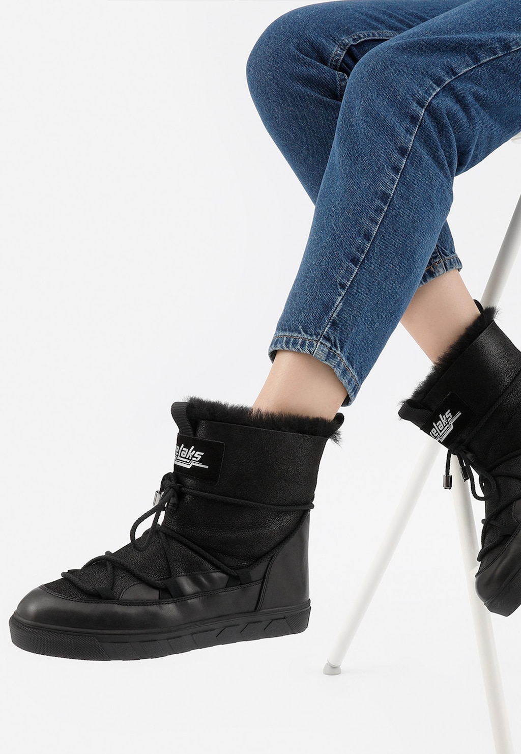 Czarne śniegowce damskie RELAKS R55103-71 z podszewką ze skóry owczej na nogach osoby siedzącej na białym stołku, widoczne z boku, zestawione z jeansami.